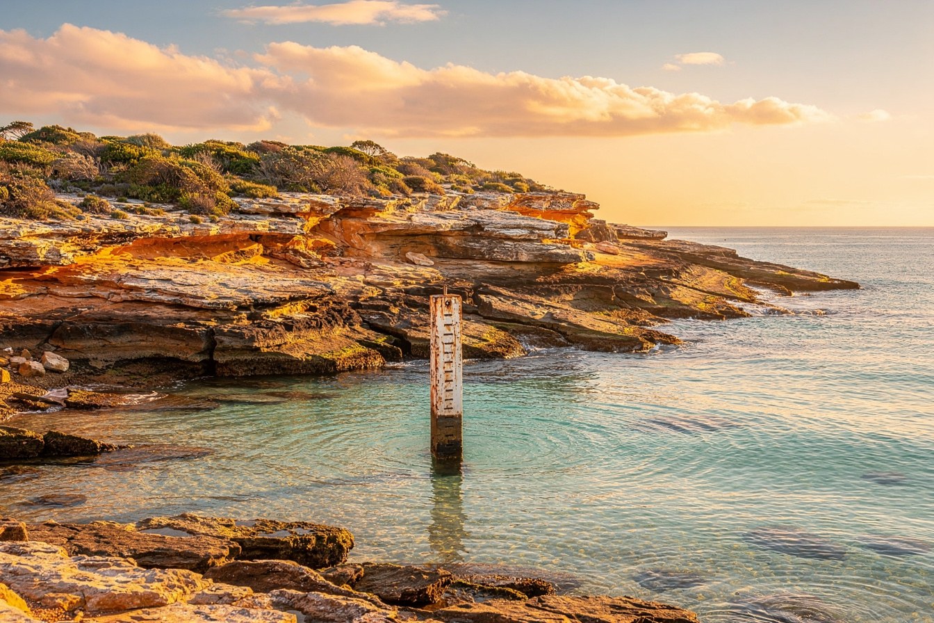 A tide gauge post in shallow coastal water at dawn, marking a line that keeps moving, standing for a record that changes after you last checked it.